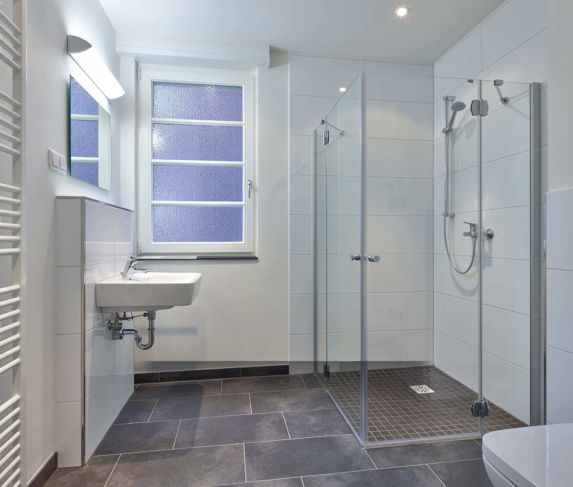 Bright modern tiled bathroom with glass shower enclosure, wall-mounted sink and dark floor tiles